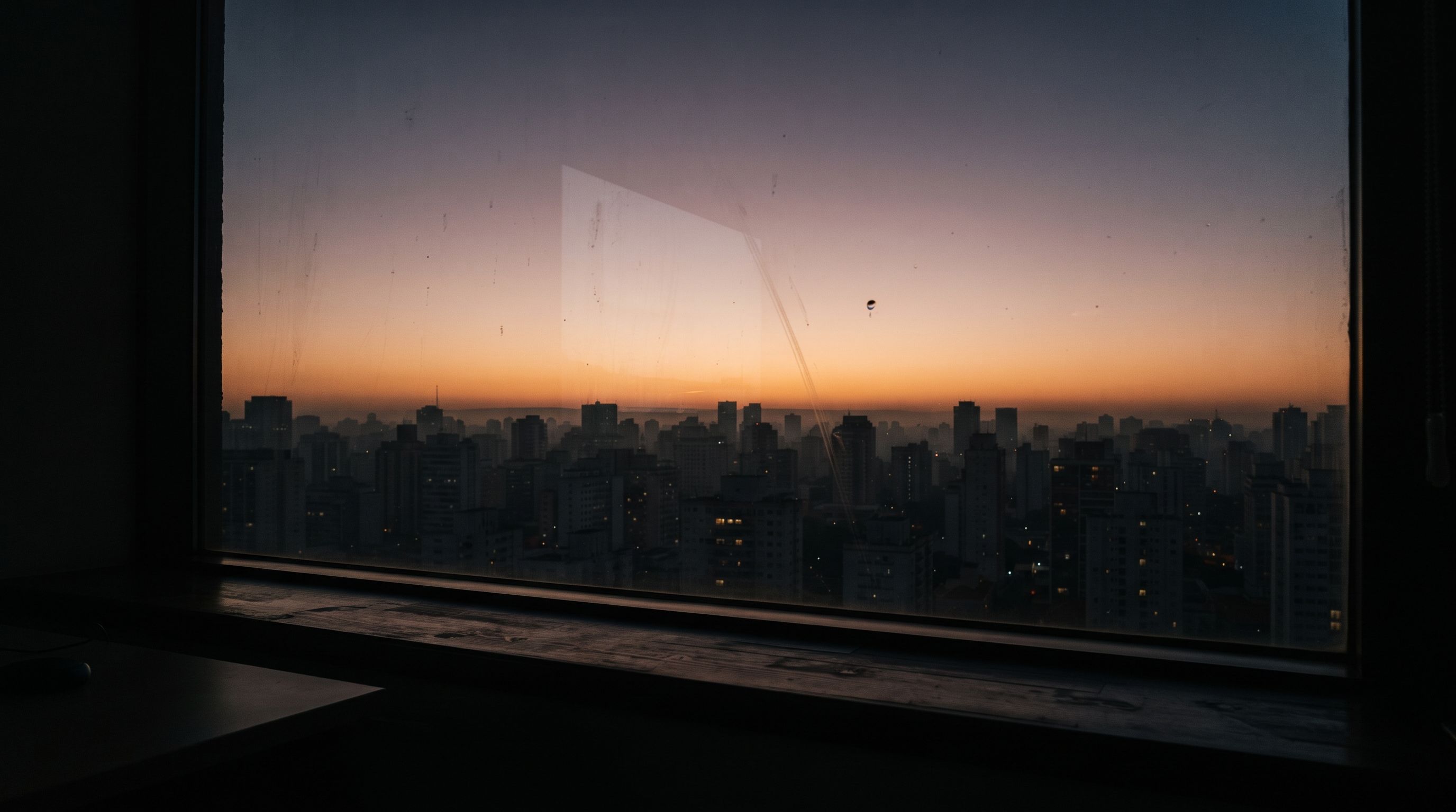 Sunrise seen from inside a dark office through a large window: a city skyline in silhouette under a band of amber light on the horizon.